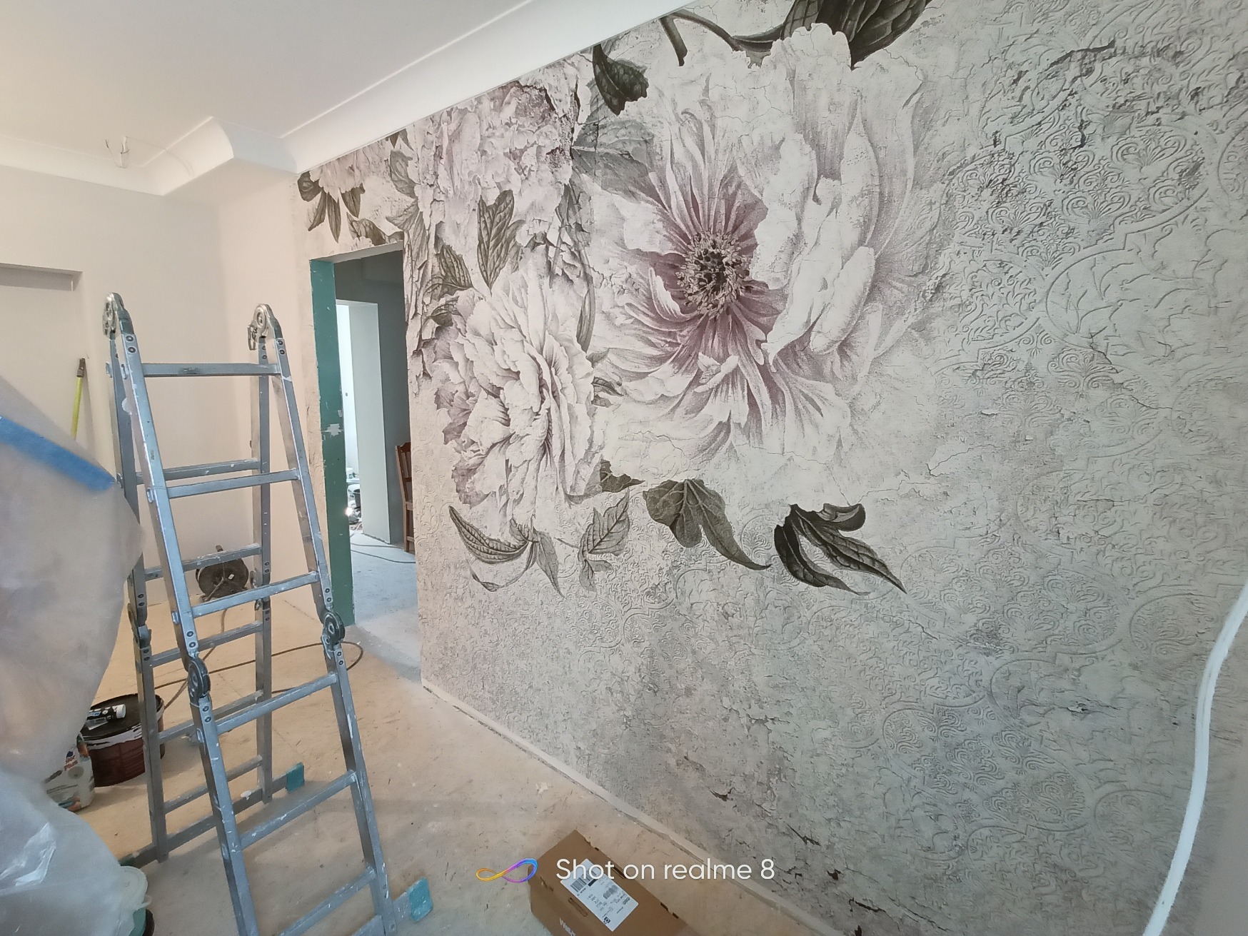 Ściana z dekoracyjną tapetą w kwiaty i ornamenty, drabina aluminiowa w tle, karton na podłodze.