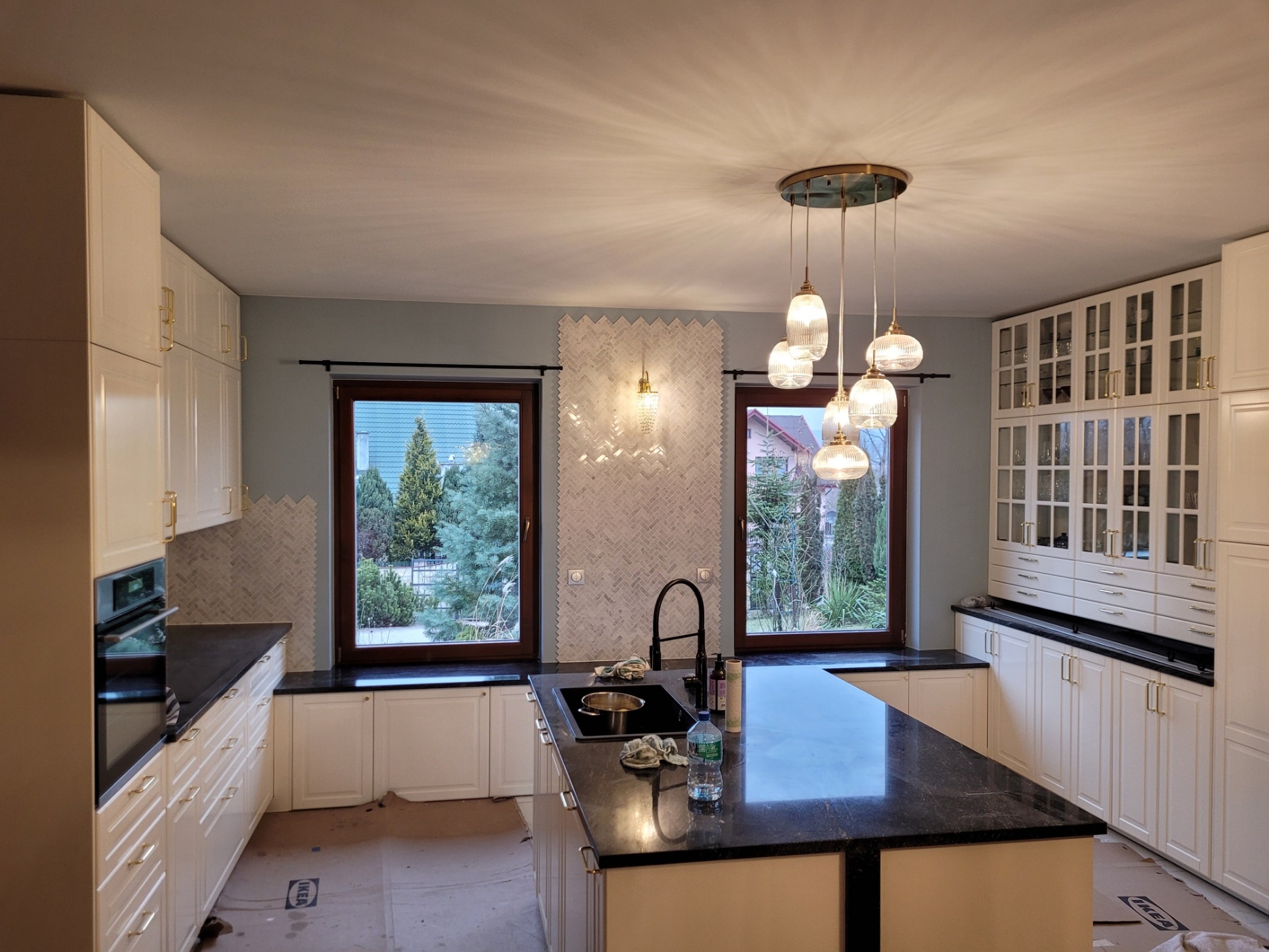 Wnętrze kuchni w trakcie prac wykończeniowych: białe szafki, czarny blat, mozaika herringbone na ścianie, wisząca lampa z kilkoma szklanymi kloszami, okna z widokiem na zieleń, podłoga...