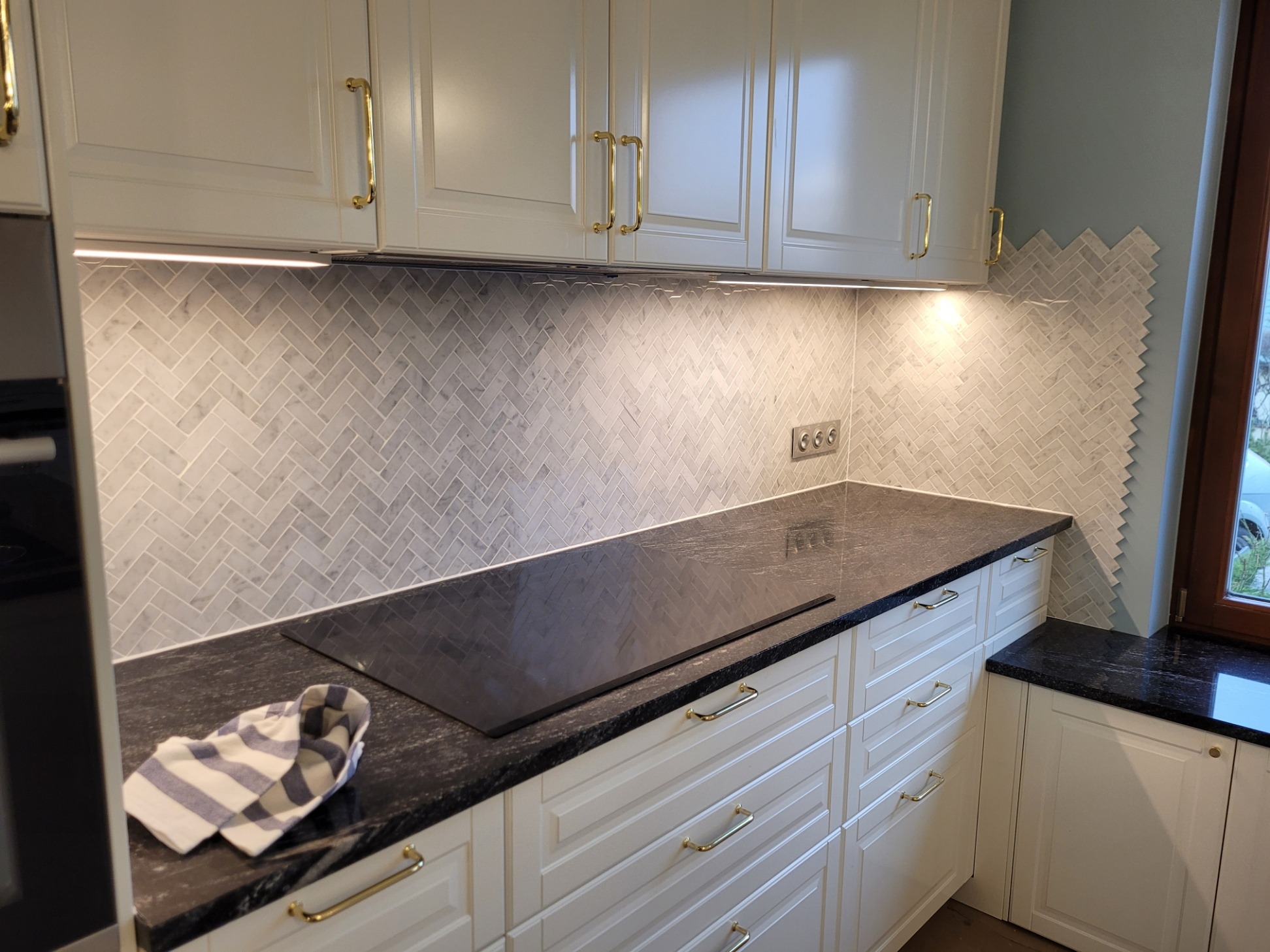 Jasna kuchnia z marmurową mozaiką ułożoną w jodełkę na ścianie, czarny kamienny blat i białe szafki z złotymi uchwytami, oświetlenie LED podszafkowe.