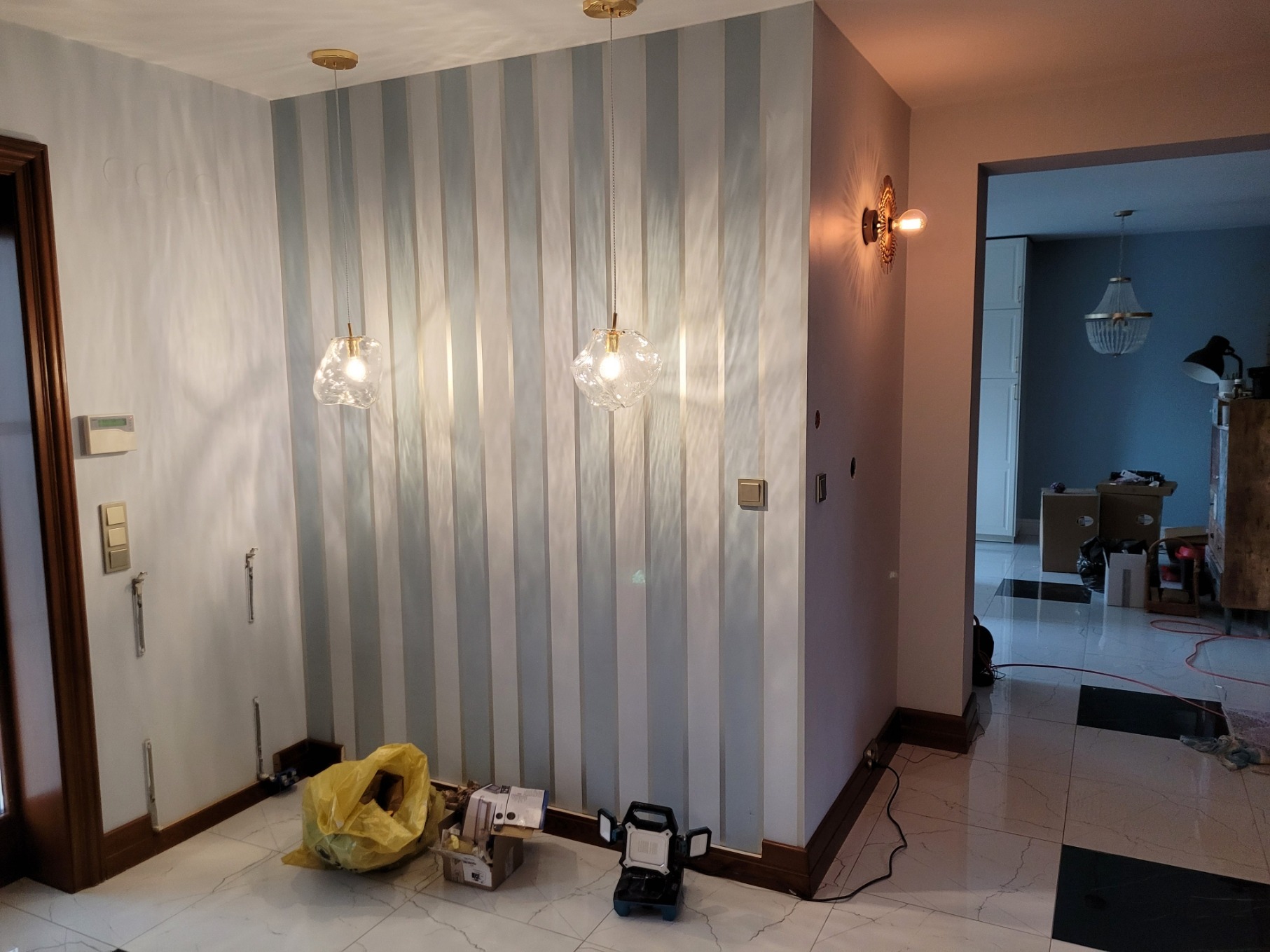 Pokój w trakcie odświeżania: ściana w biało-szare pasy z wiszącymi lampami o nieregularnych kloszach, widoczne ślady po gniazdkach, na podłodze torba z narzędziami i lampa robocza.