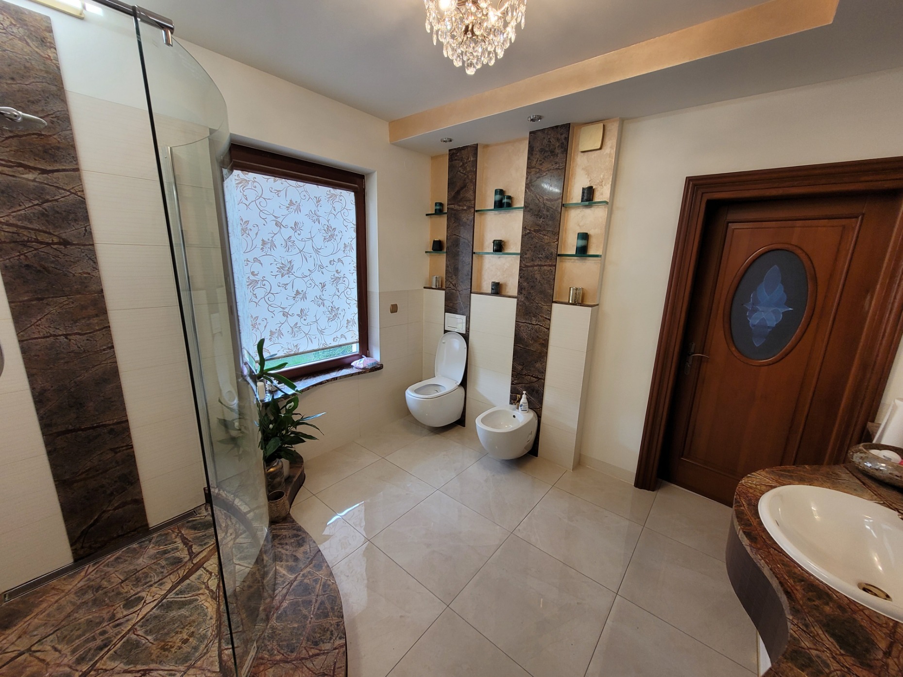 Jasna łazienka z kabiną prysznicową z marmurowym wykończeniem, toaletą, bidetem i umywalką z blatem z marmuru. Na ścianie półki z dekoracjami. Okno z roletą w kwiaty.