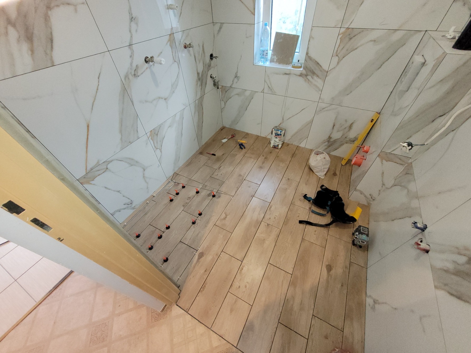 Łazienka w trakcie układania płytek podłogowych imitujących drewno, z widocznymi klipsami poziomującymi i białymi płytkami marmurkowymi na ścianach, częściowo wykończonych, z niepodłączoną armaturą.