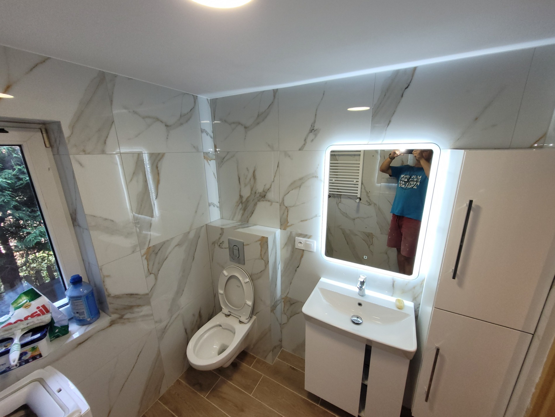 Wykończona łazienka z toaletą podwieszaną, umywalką z szafką, lustrem z podświetleniem LED i płytkami imitującymi marmur, widoczne środki czystości na parapecie okna.