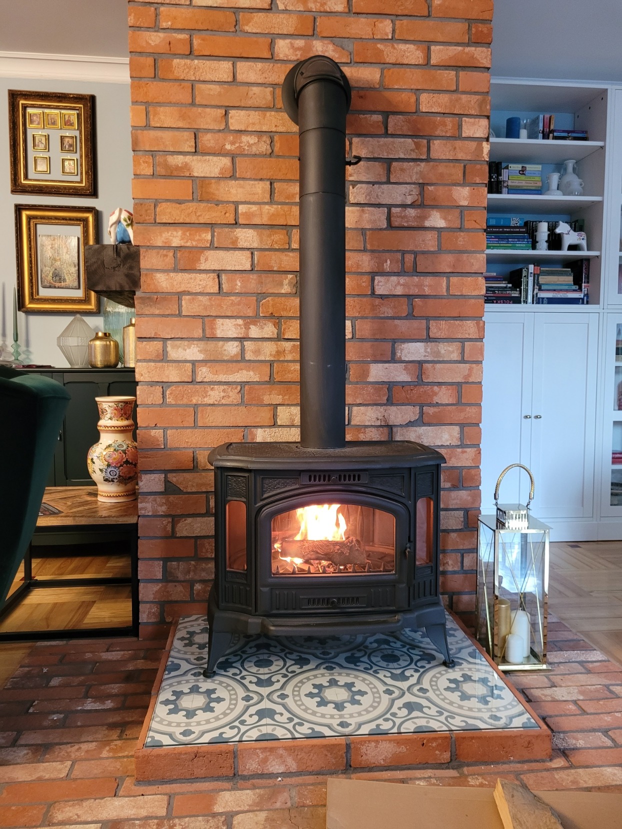 Ognisko w żeliwnym piecu kominkowym z rurą odprowadzającą dym, obudowane cegłą, na dekoracyjnych płytkach podłogowych, z lampionem obok, w tle półki z książkami.