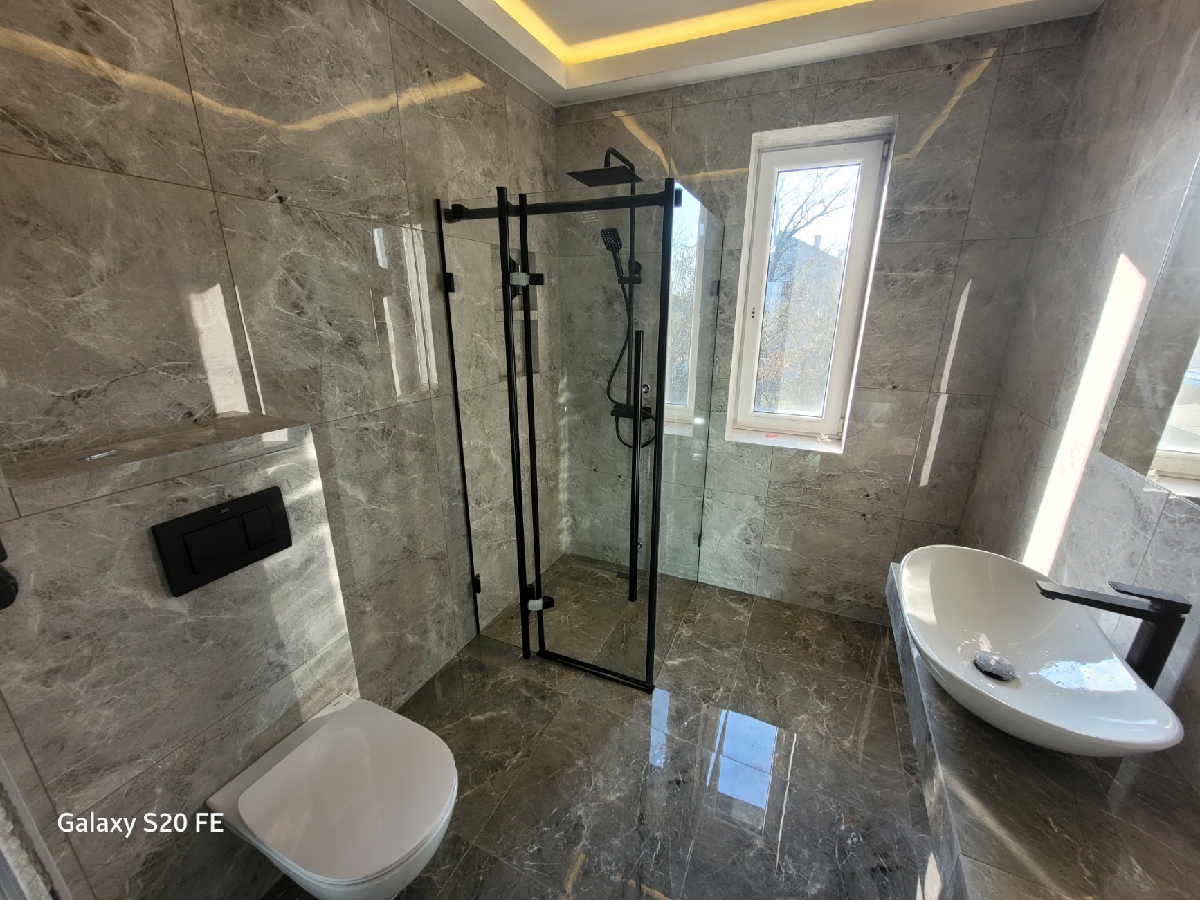 Nowoczesna łazienka z szarymi marmurkowymi płytkami, kabiną prysznicową typu walk-in z czarnymi profilami, wiszącą miską WC z czarnym przyciskiem spłukującym i owalną umywalką nablatową.