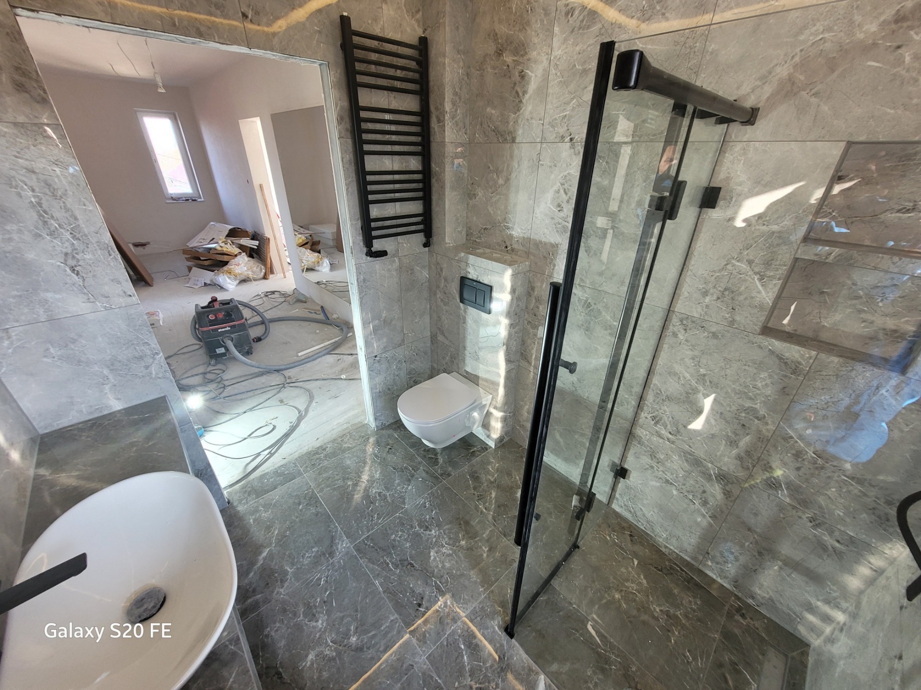 Wnętrze łazienki w trakcie prac wykończeniowych, widoczna kabina prysznicowa z czarnymi profilami, umywalka nablatowa, toaleta podwieszana i grzejnik drabinkowy na tle marmurowych płytek, w tle...