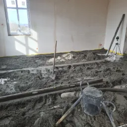 Wnętrze w trakcie wylewania posadzki: widoczna szlichta cementowa, rury ogrzewania podłogowego, poziomica, łopata i niwelator laserowy na statywie.