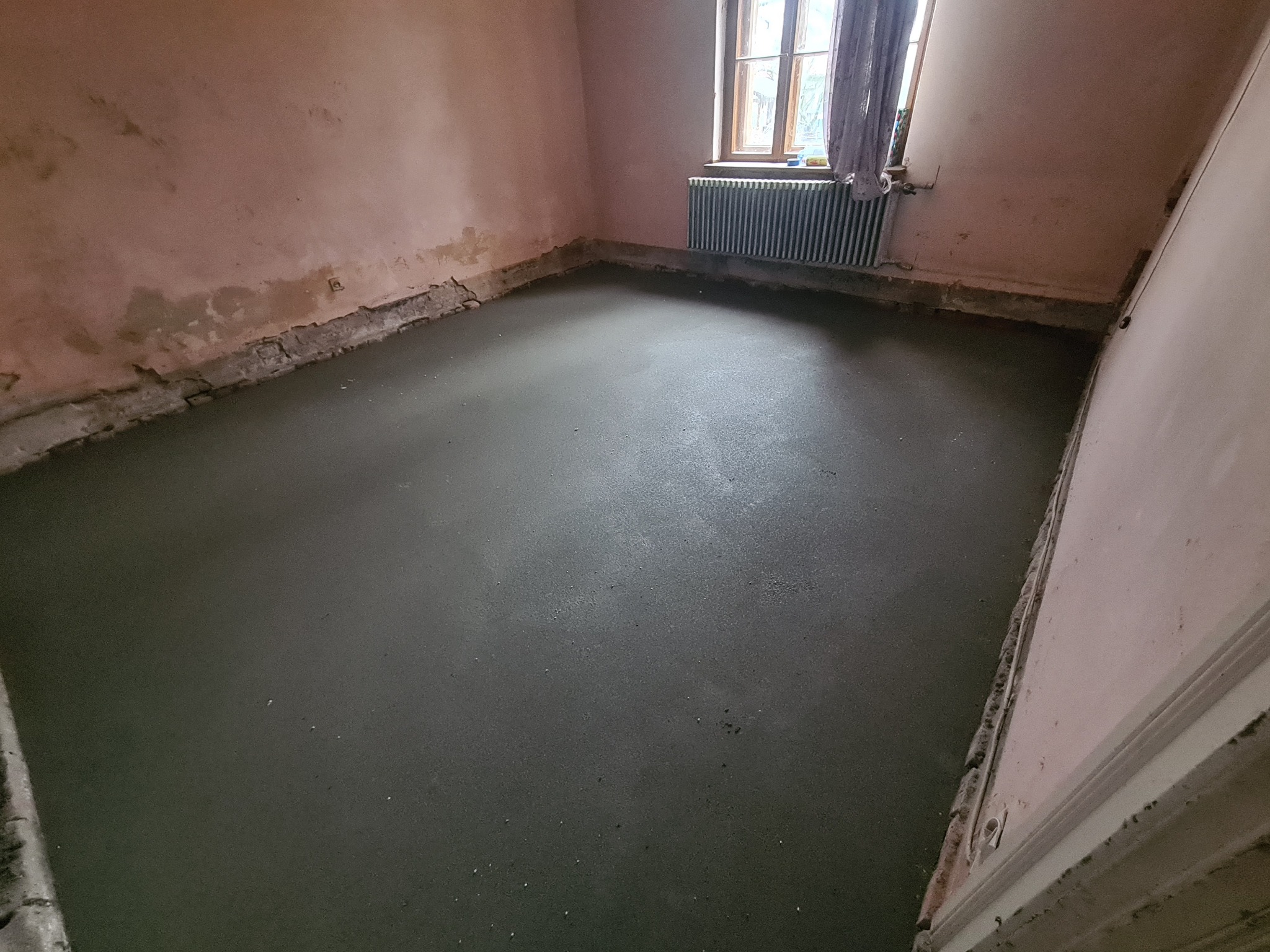 Świeżo wylana, gładka posadzka jastrychowa w pokoju z oknem i starym grzejnikiem, ściany w trakcie remontu, widoczne ślady po wcześniejszych warstwach wykończeniowych.