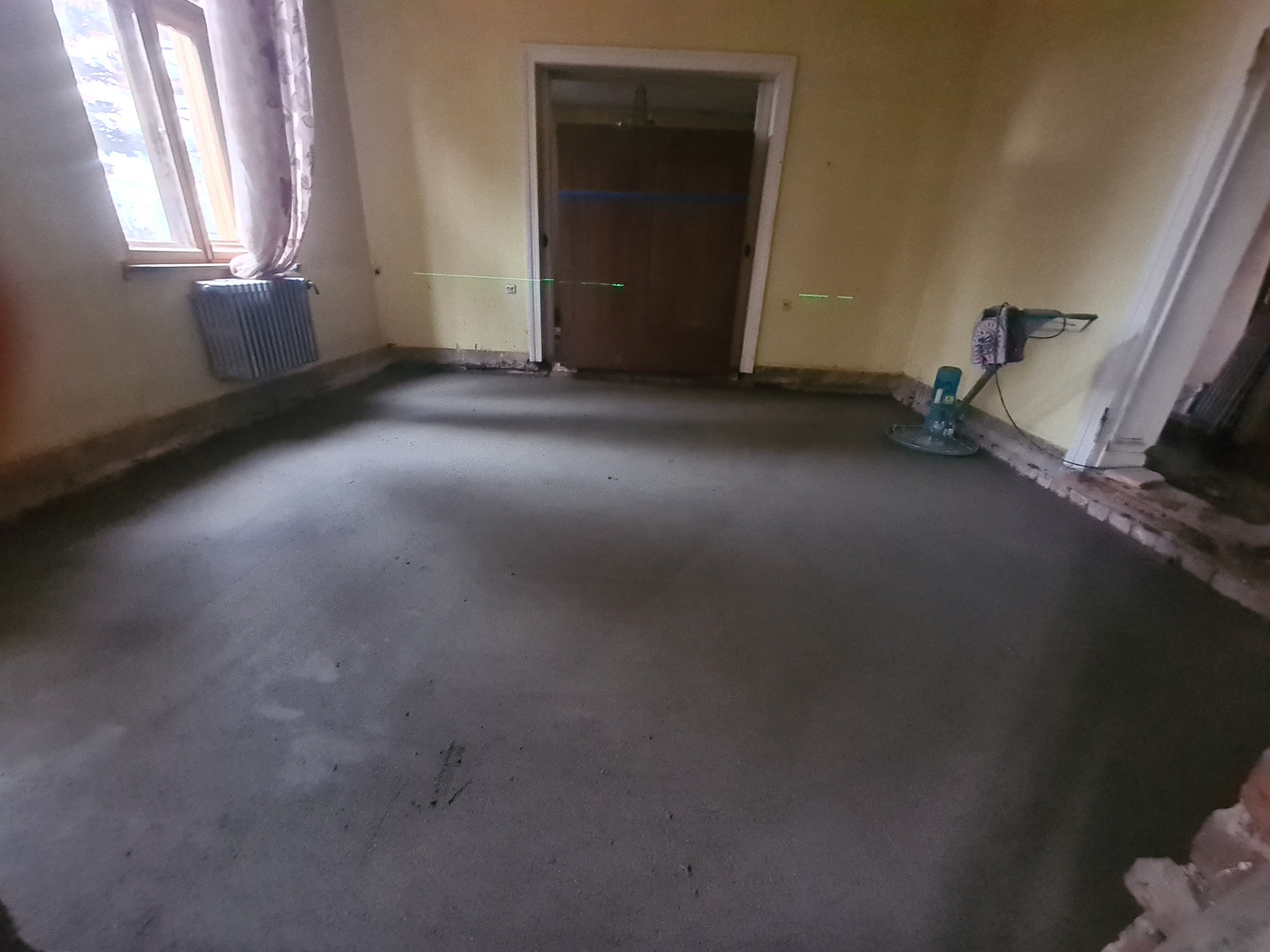 Świeżo wylana, szara posadzka w pomieszczeniu z widocznym laserowym niwelatorem, grzejnikiem i szlifierką do betonu.