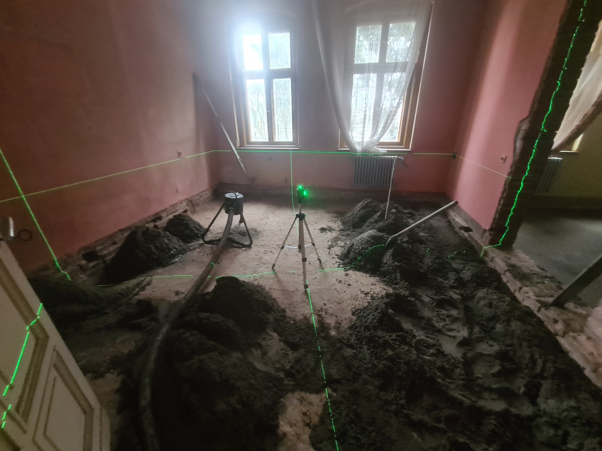 Przygotowanie podłoża pod posadzkę: nierównomiernie rozłożona masa wyrównująca w pokoju z dwoma oknami, poziomica laserowa wyznacza poziom na ścianach.