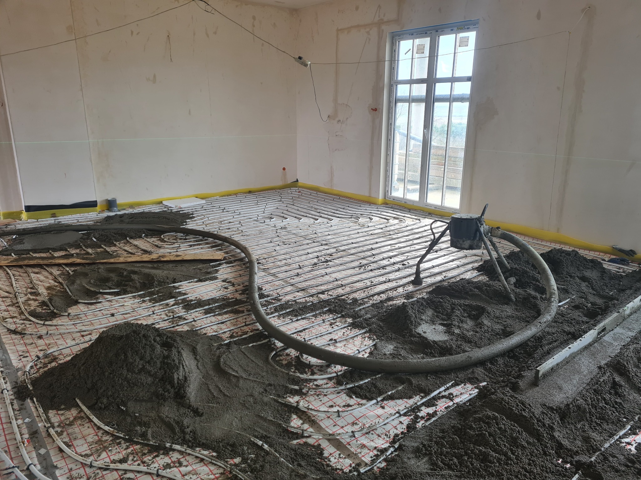 Wnętrze w trakcie wylewania podkładu podłogowego z ogrzewaniem podłogowym, widoczne rury grzewcze, warstwa izolacyjna przy ścianach, oraz hałdy mokrej zaprawy i poziomica.