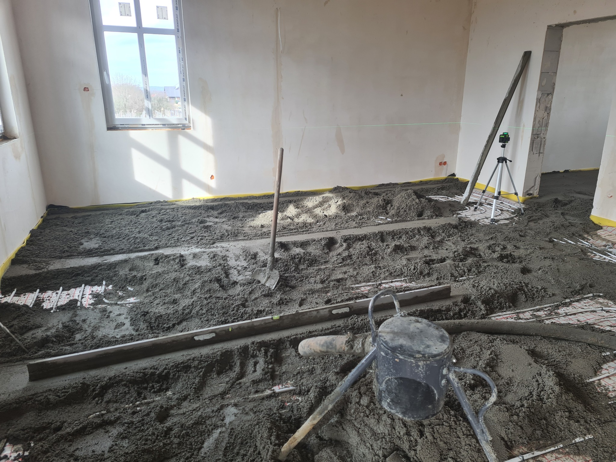 Wnętrze w trakcie wylewania posadzki: widoczna szlichta cementowa, rury ogrzewania podłogowego, poziomica, łopata i niwelator laserowy na statywie.