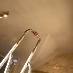 Metalowa drabina stojąca na tle świeżo pomalowanego sufitu z widocznymi śladami po taśmie malarskiej, fragment ściany wykończonej płytkami ceramicznymi w tle. Oświetlenie punktowe na suficie.