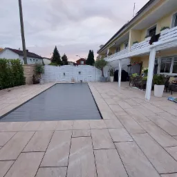 Zewnętrzny taras z jasnych, prostokątnych płytek ceramicznych otaczający zakryty basen z automatyczną roletą, widok na biały płot i zabudowę domu z balkonami.