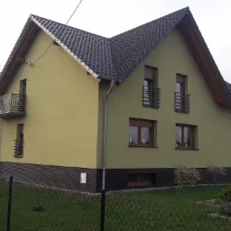 Dwupiętrowy dom z ciemną dachówką, zieloną elewacją i małymi balkonami z metalowymi balustradami, widoczny zza ogrodzenia.