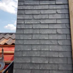 Fragment dachu krytego łupkiem, widoczne otwory wentylacyjne i fragment sąsiedniego dachu z dachówki ceramicznej oraz narzędzie do cięcia kamienia.