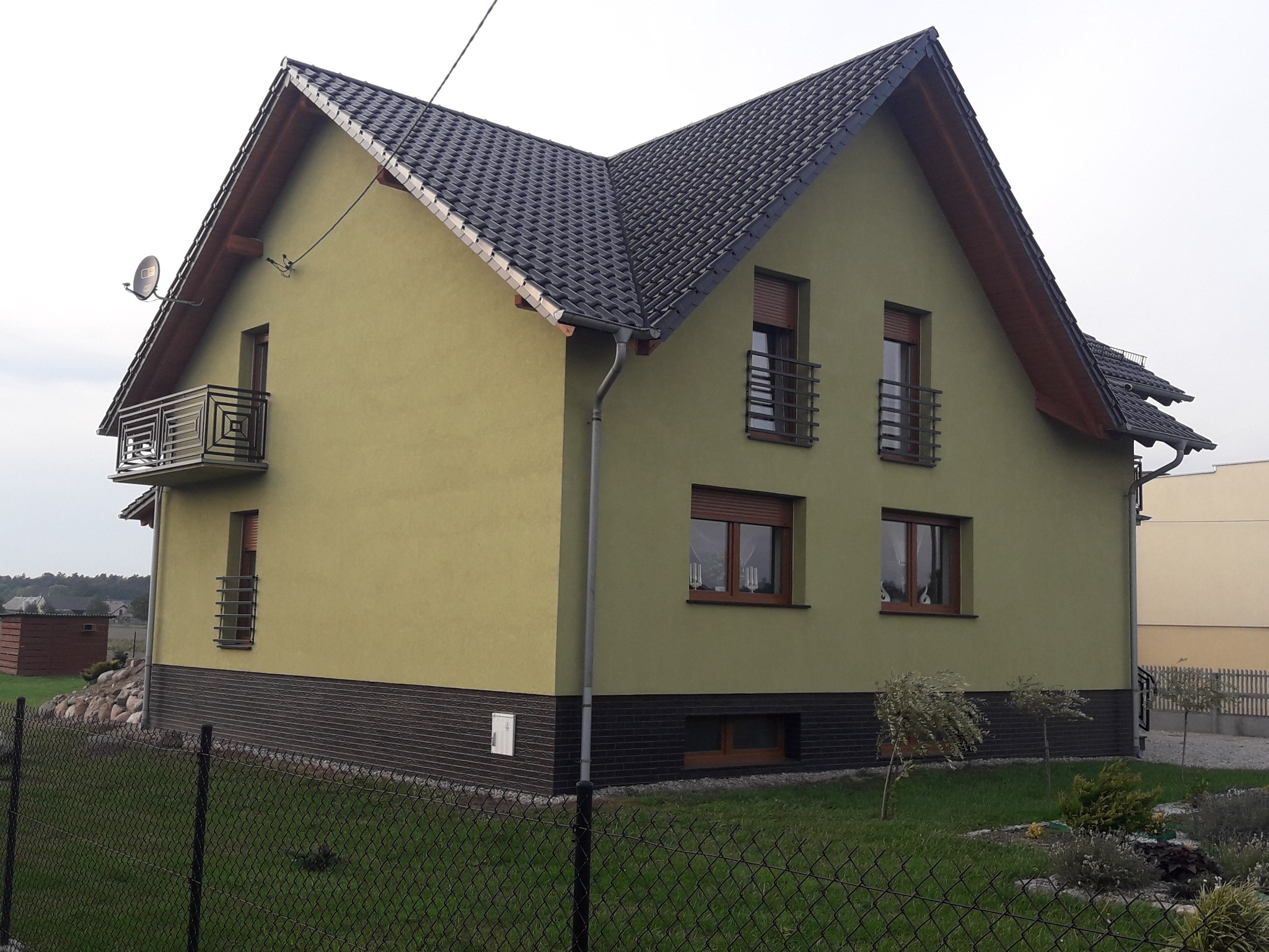 Dwupiętrowy dom z ciemną dachówką, zieloną elewacją i małymi balkonami z metalowymi balustradami, widoczny zza ogrodzenia.