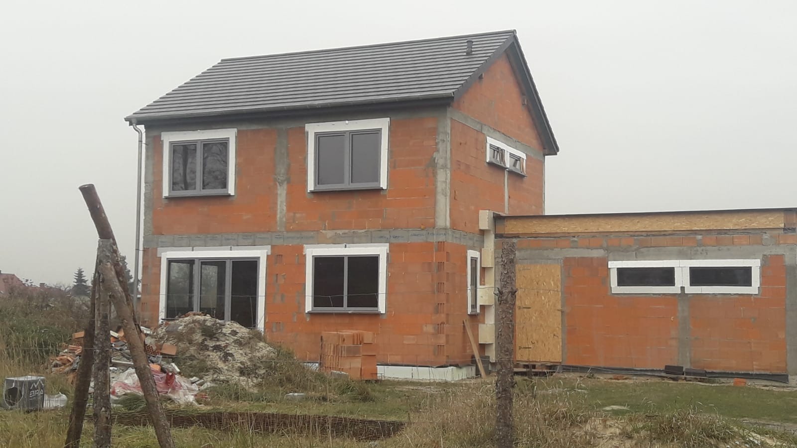 Surowy stan dwukondygnacyjnego domu z szarą dachówką i szarą stolarką okienną, widoczne cegły i elementy konstrukcyjne, teren budowy z gruzem i stertą piasku.