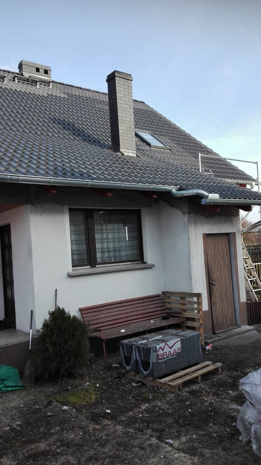 Dom z nowym dachem pokrytym szarą dachówką Braas, widoczne rynny, komin z cegły, okno dachowe i rusztowanie, na ziemi paleta z dachówkami Braas.