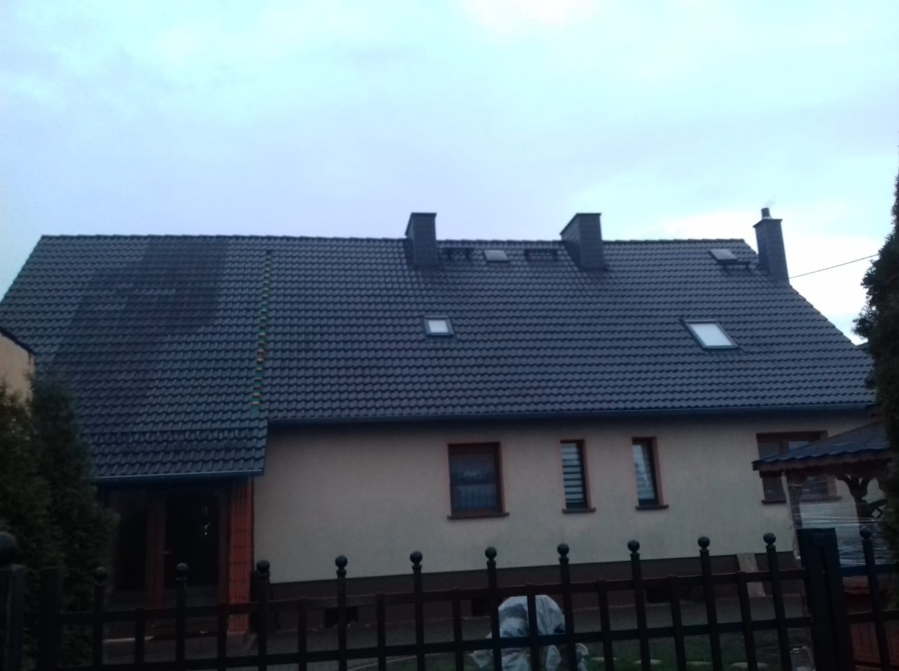 Dom jednorodzinny z ciemną dachówką, widoczne kominy, okna dachowe i fragment zabrudzonego dachu z mchem po lewej stronie.