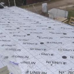 Membrana dachowa UNILIN z widocznymi mocowaniami, na tle częściowo widoczny budynek i elementy konstrukcyjne dachu, robocze rękawice leżą na powierzchni membrany.