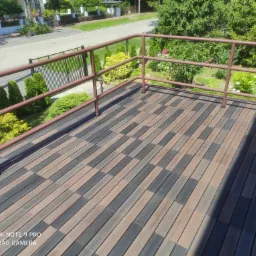 Balkon z brązową balustradą i podłogą z desek kompozytowych w odcieniach brązu i szarości, widok na zadbany ogród i ulicę.