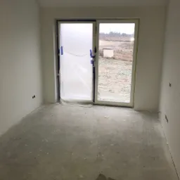 Surowe, puste pomieszczenie w trakcie prac wykończeniowych; ściany pokryte świeżą gładzią, okno zabezpieczone folią malarską, widoczne puszki elektryczne, podłoga betonowa z resztkami materiałów...