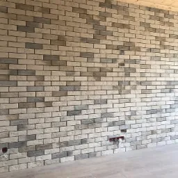 Ściana z dekoracyjnej cegły w odcieniach szarości i beżu, widoczne puszki elektryczne bez osprzętu, drewniany sufit, jasna podłoga.