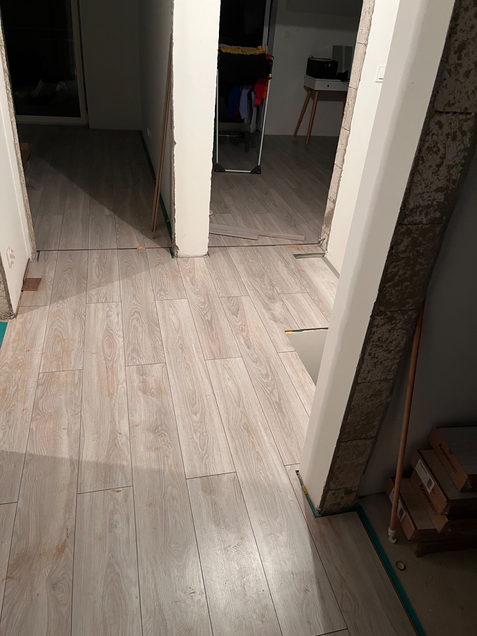 Ułożona podłoga z paneli imitujących drewno w trakcie prac wykończeniowych, widoczne nierówne krawędzie przy futrynach i szczeliny dylatacyjne zabezpieczone zieloną taśmą, w tle częściowo widoczne...