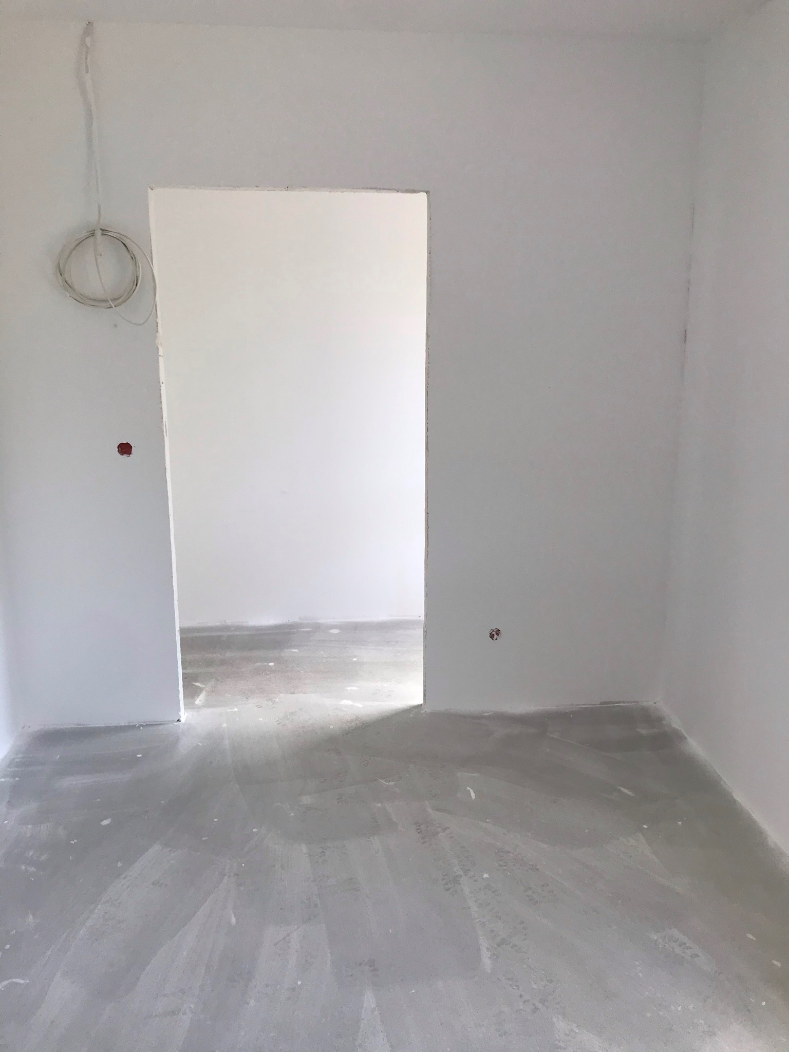 Przygotowane do malowania, białe ściany w pustym pokoju, widoczne ślady szpachlowania na podłodze i wystające przewody elektryczne.