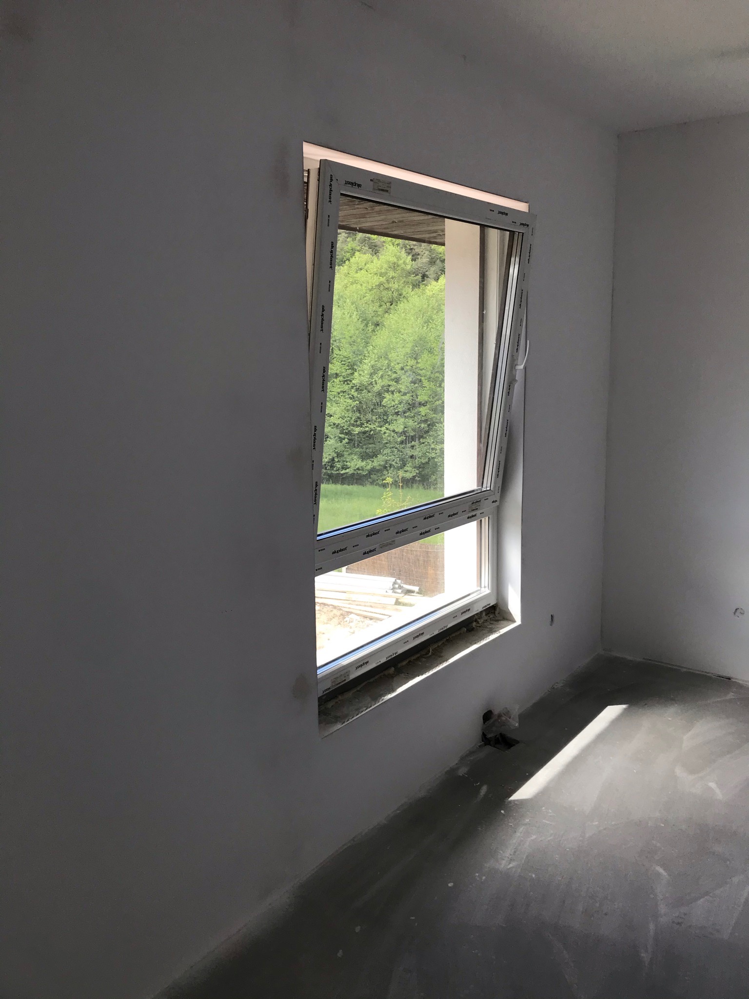 Wnętrze w stanie surowym z uchylonym oknem PCV marki Aluplast, przez które widać zieleń drzew. Podłoga betonowa, ściany świeżo otynkowane, widoczne ślady po instalacji elektrycznej.