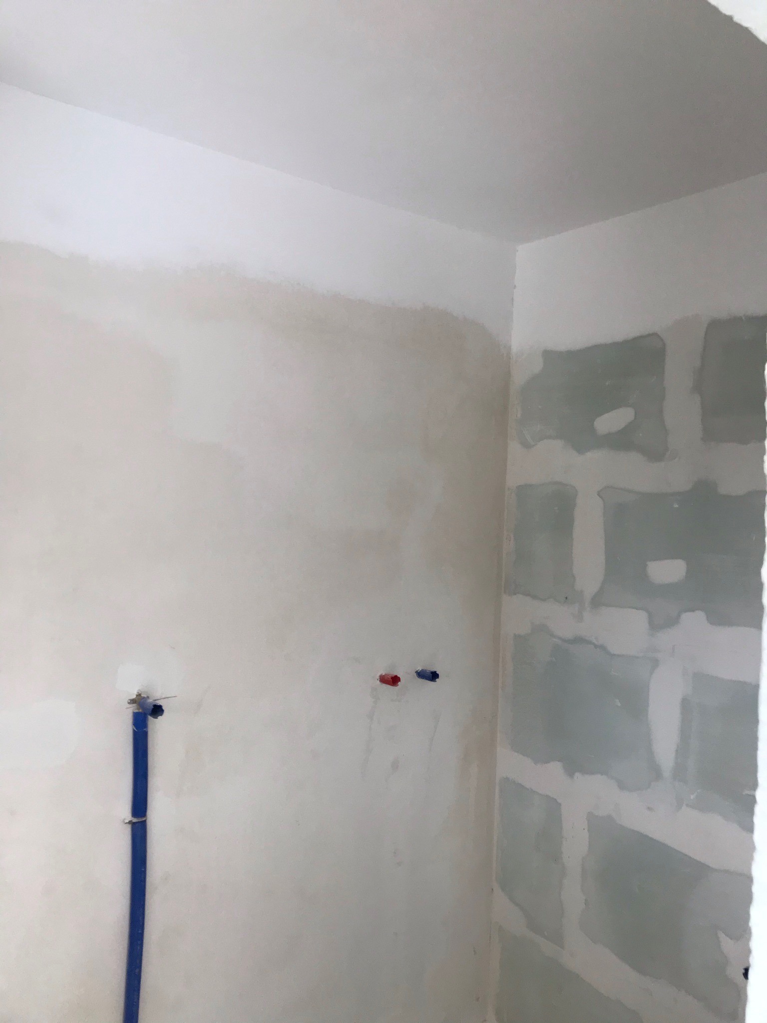 Wnętrze w trakcie prac wykończeniowych: ściana z fragmentami nałożonej gładzi, widoczne rury instalacji wodnej, ściana z płyt kartonowo-gipsowych z widocznymi łączeniami.