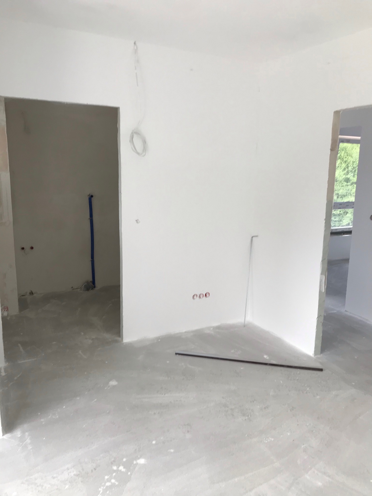 Surowe wnętrze w trakcie prac wykończeniowych: białe ściany, szary betonowy podkład, widoczne instalacje elektryczne i hydrauliczne, przygotowane do dalszych prac.