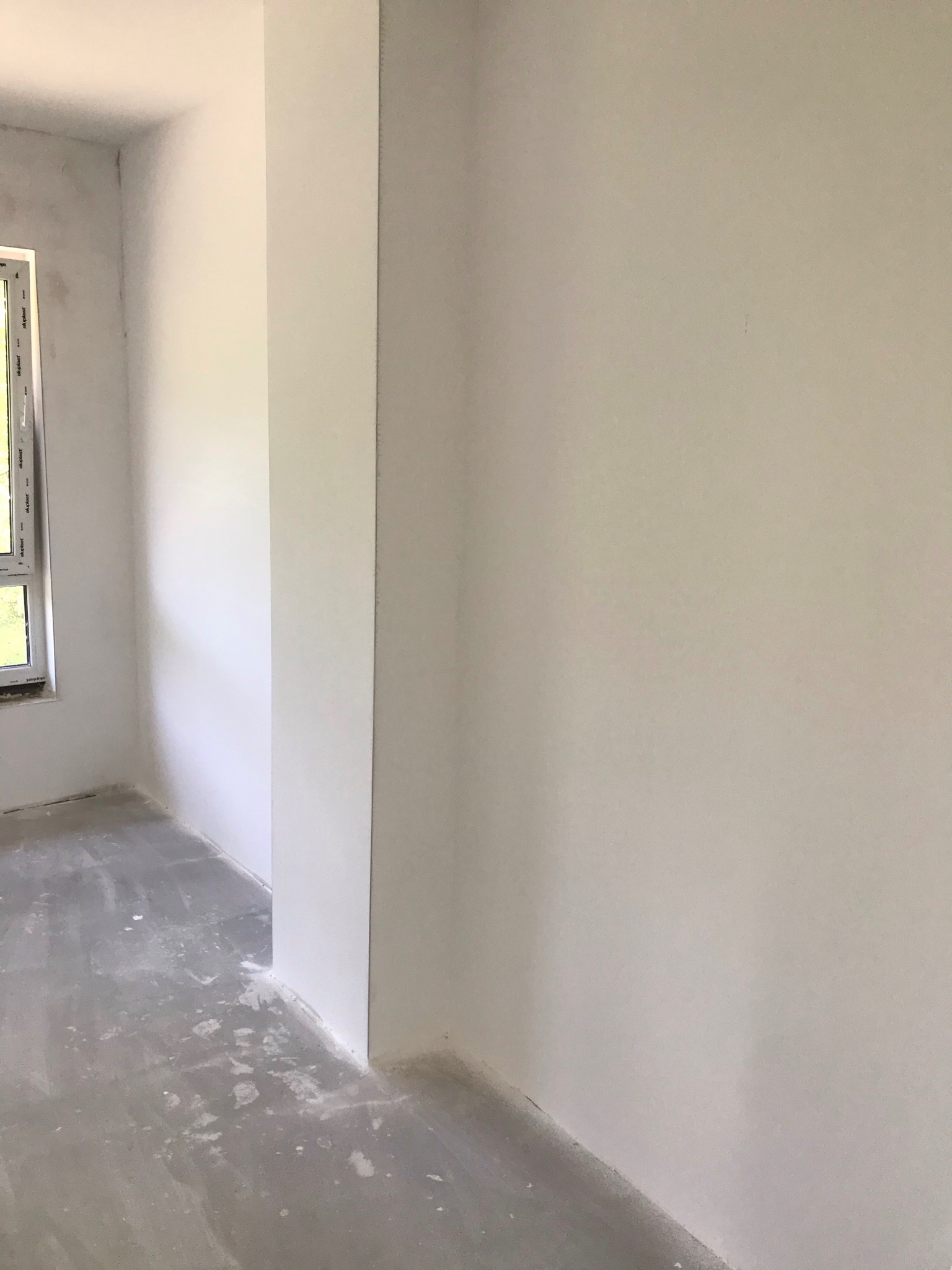 Surowe wnętrze pokoju po świeżo wykonanych pracach: białe ściany, szary betonowy podkład z resztkami zaprawy, okno z widokiem na zieleń.