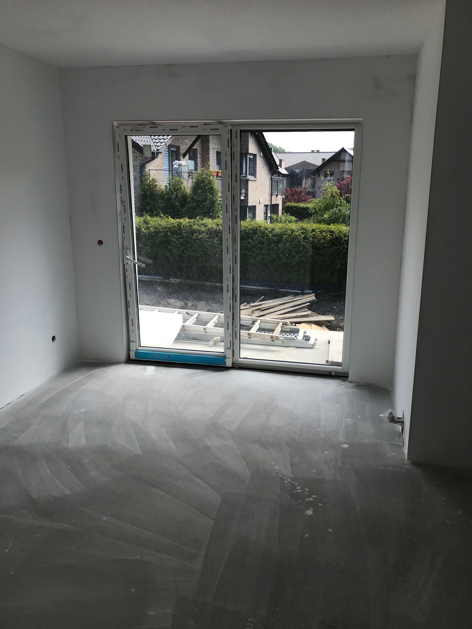 Surowe wnętrze pokoju z przeszklonymi drzwiami balkonowymi; widoczne przygotowane ściany z widocznymi gniazdkami, wylewka na podłodze oraz drabina i deski za oknem.