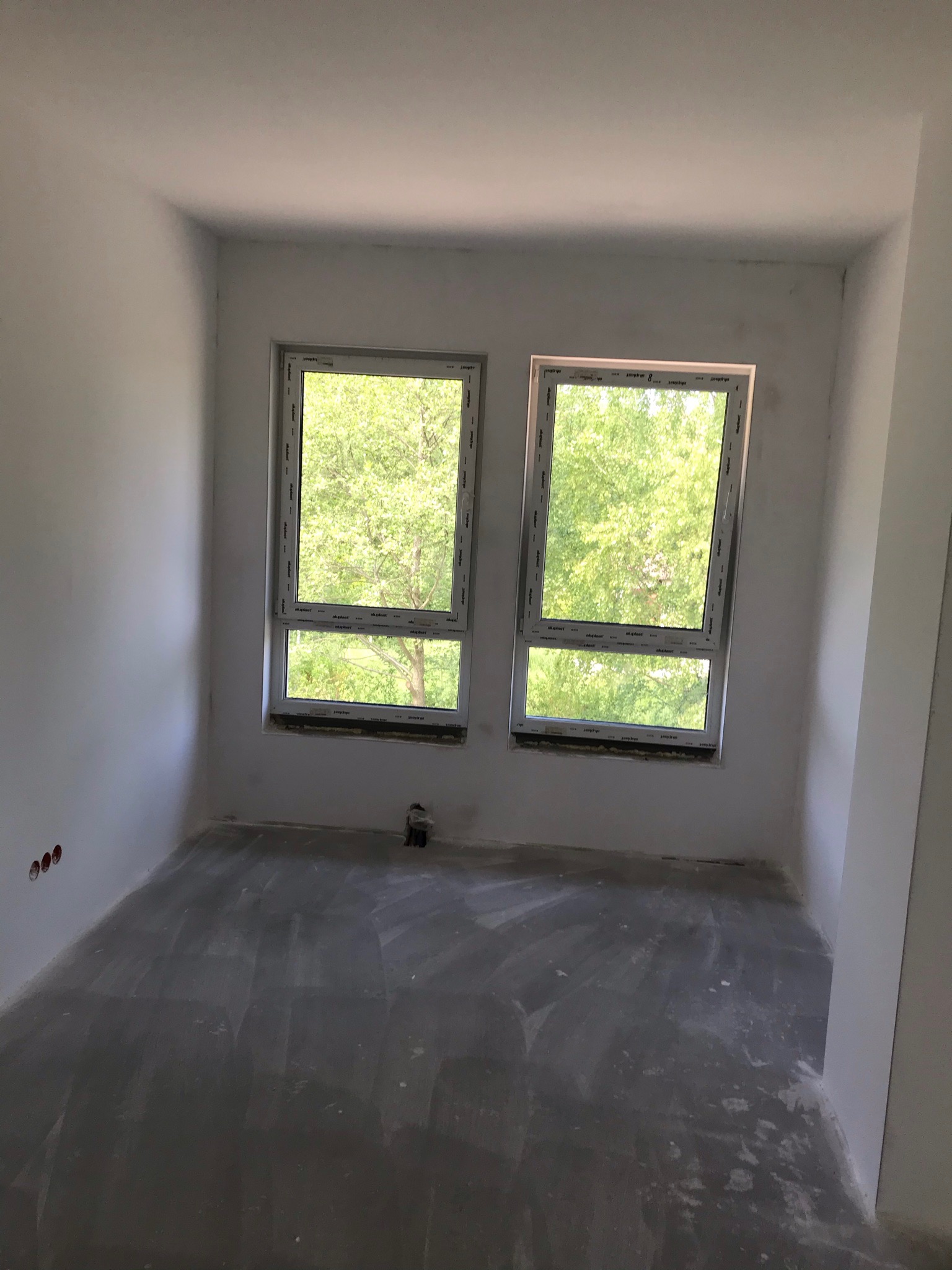 Surowe wnętrze pokoju po wylewce podłogi, z dwoma białymi oknami z widokiem na zieleń, ściany przygotowane do malowania, widoczne gniazdka elektryczne.