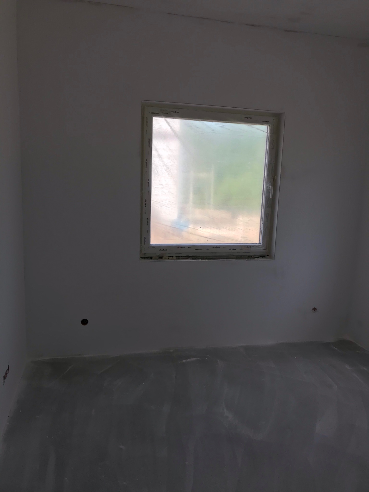 Pokój w trakcie prac wykończeniowych: świeżo nałożona gładź na ścianach i podłodze, okno z folią ochronną.