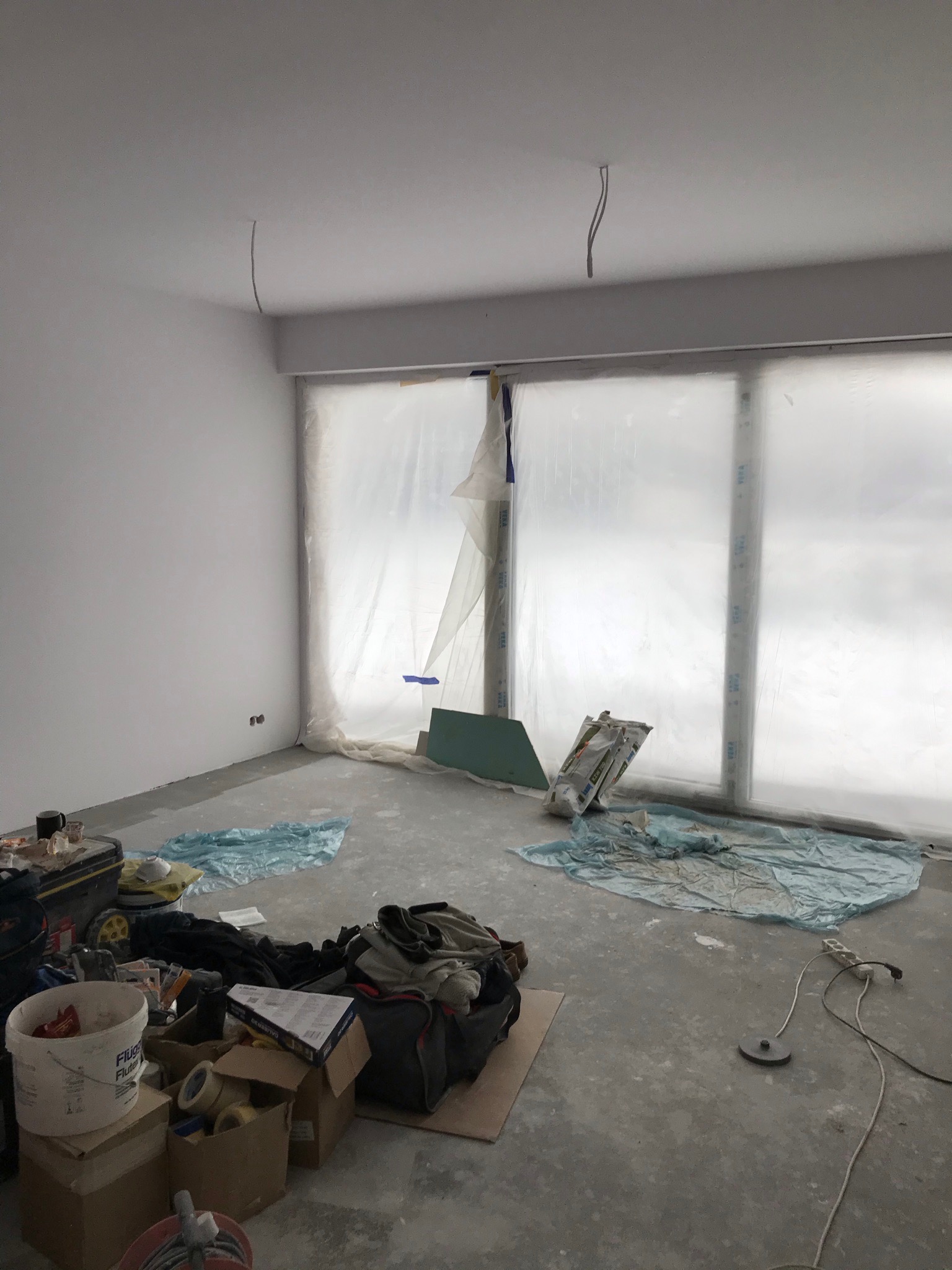 Pokój w trakcie remontu: ściany pomalowane na biało, okna zabezpieczone folią malarską, widoczne narzędzia i materiały budowlane, surowy beton na podłodze, wystające kable z sufitu.
