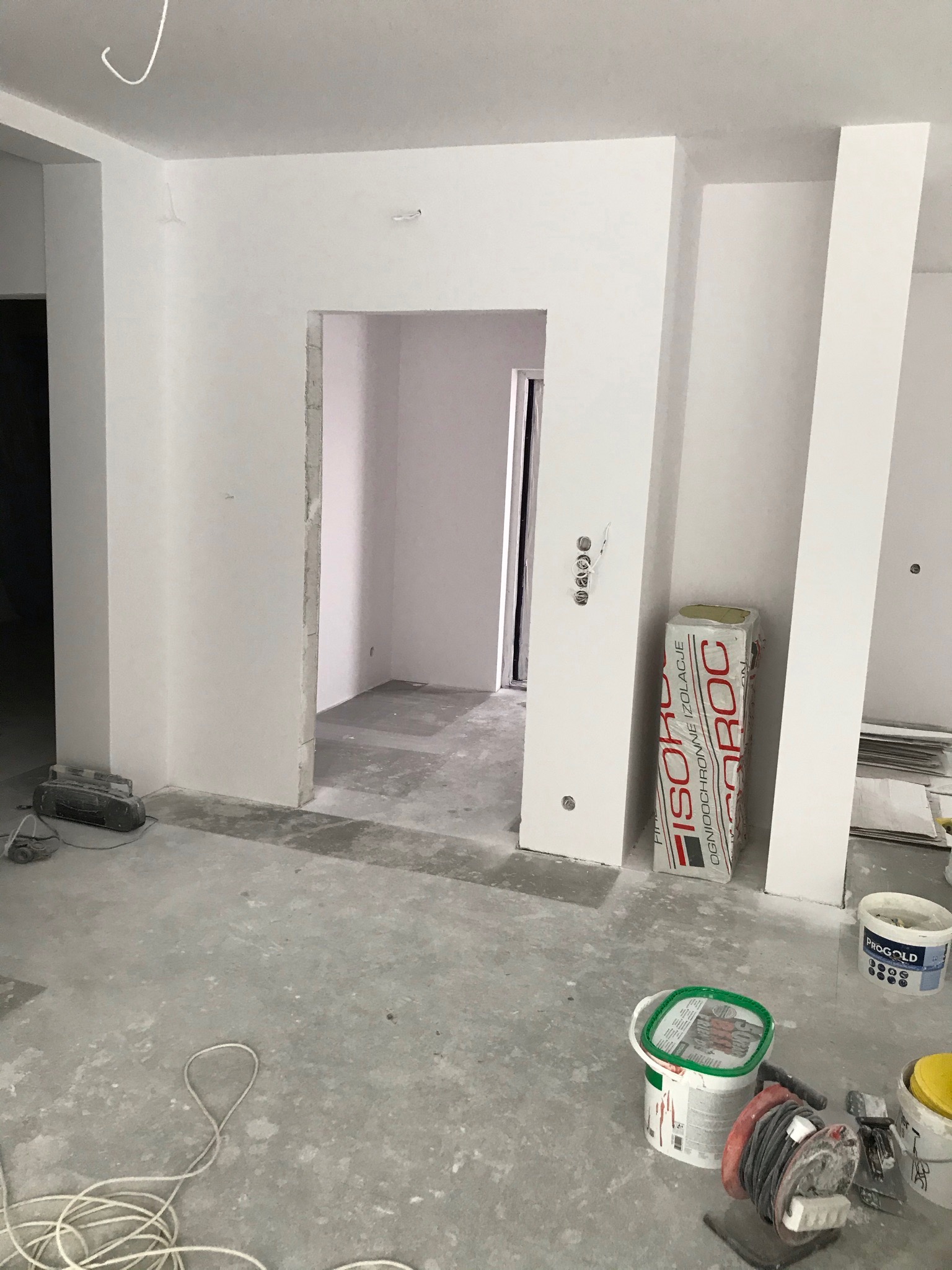Wnętrze w trakcie remontu: surowe ściany pomalowane na biało, widoczne narzędzia malarskie, puszki z farbą, płyty izolacyjne Isover, oraz niedokończone obramowanie drzwi.