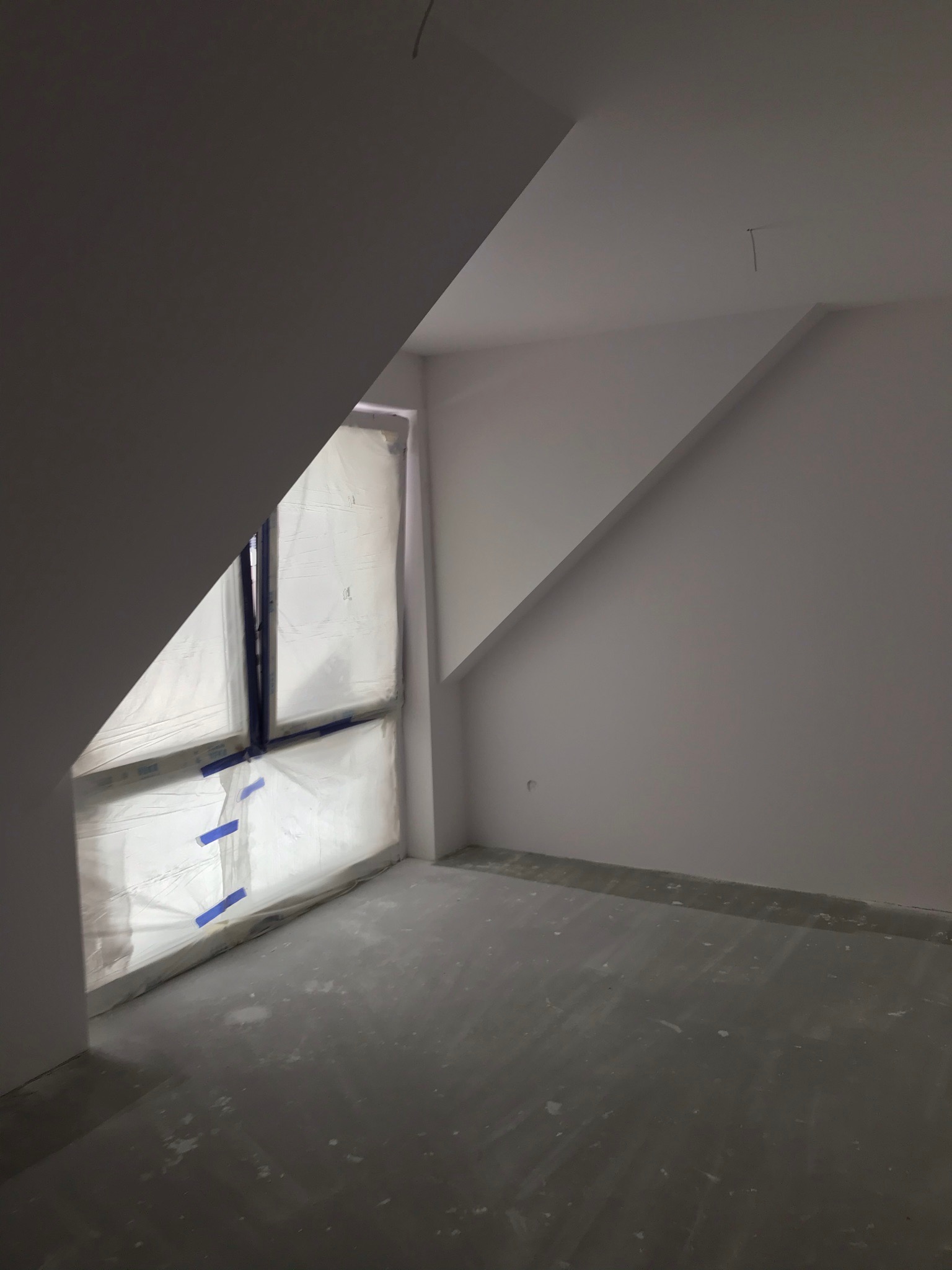 Poddasze w trakcie prac przygotowawczych: okno zabezpieczone folią i taśmą malarską, surowe ściany i podłoga przed wykończeniem.