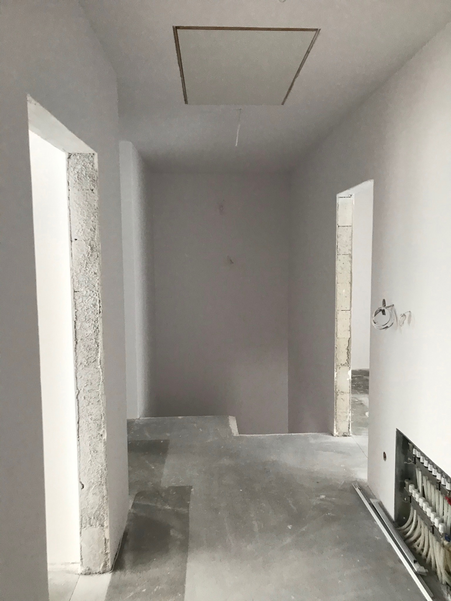 Korytarz w trakcie remontu: surowe ściany po gładziowaniu, widoczne instalacje elektryczne i hydrauliczne, przygotowane do dalszych prac wykończeniowych, otwór rewizyjny w suficie.