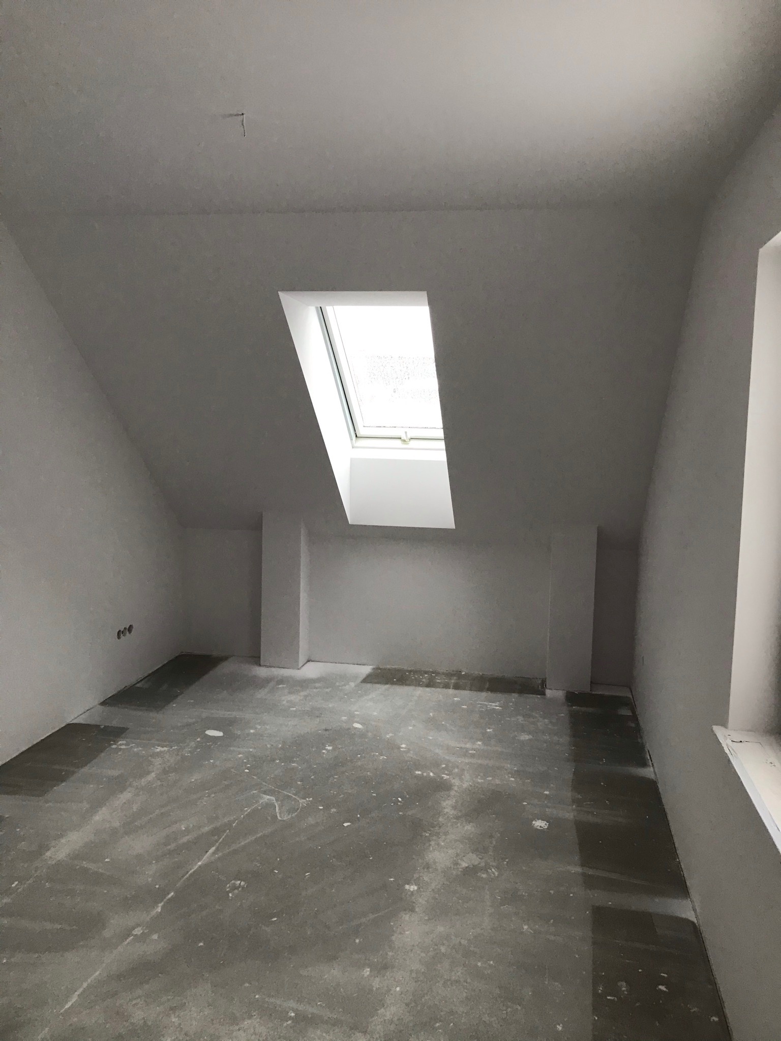 Poddasze w trakcie prac wykończeniowych, widoczne ślady gładzi na ścianach i suficie, okno dachowe wpuszczające naturalne światło, zabezpieczona podłoga, przygotowane gniazdka elektryczne.