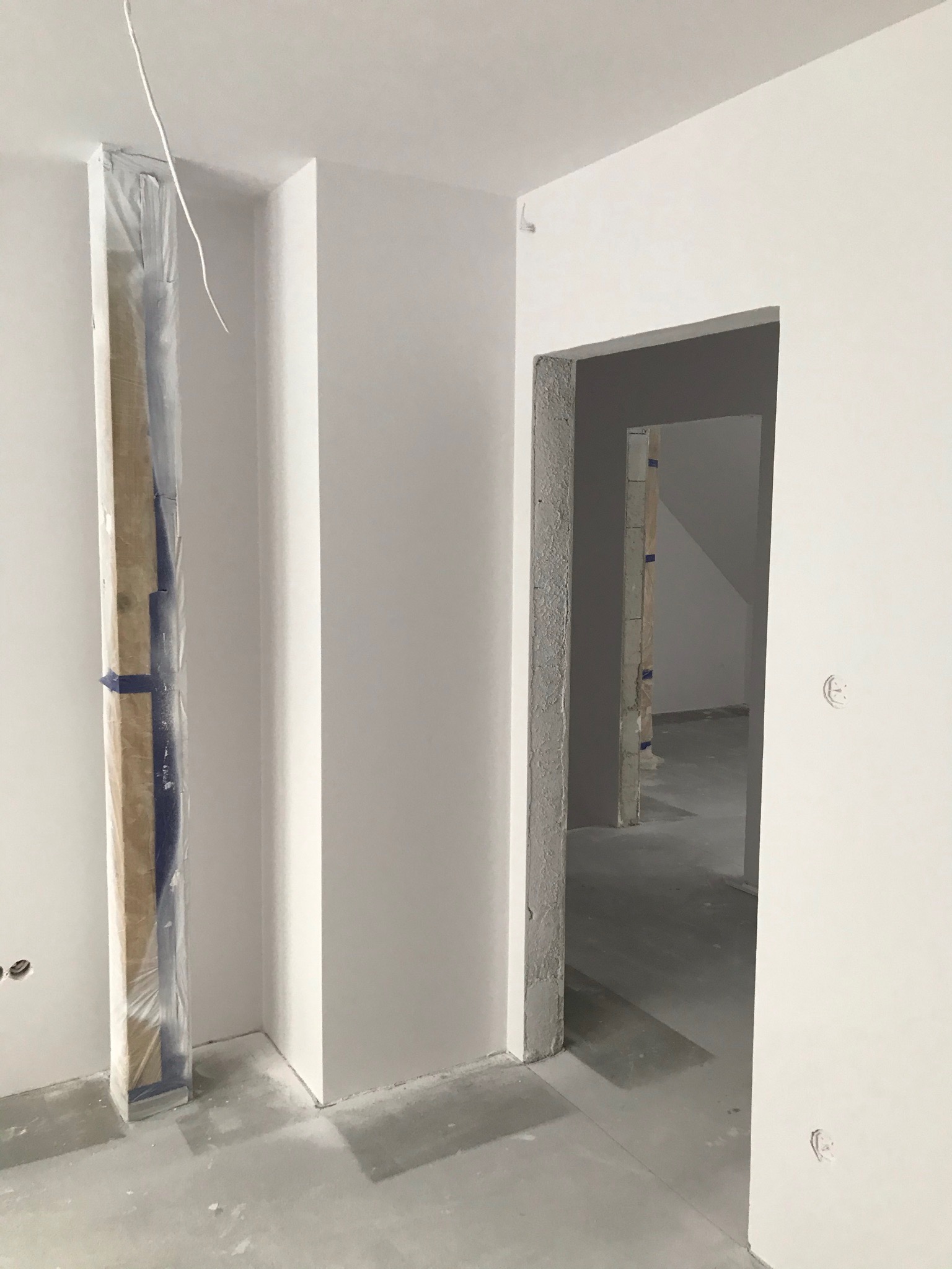 Wnętrze w trakcie prac wykończeniowych: świeżo położone białe gładzie na ścianach, surowy betonowy oścież, zabezpieczone folią profile narożnikowe przy ścianie.