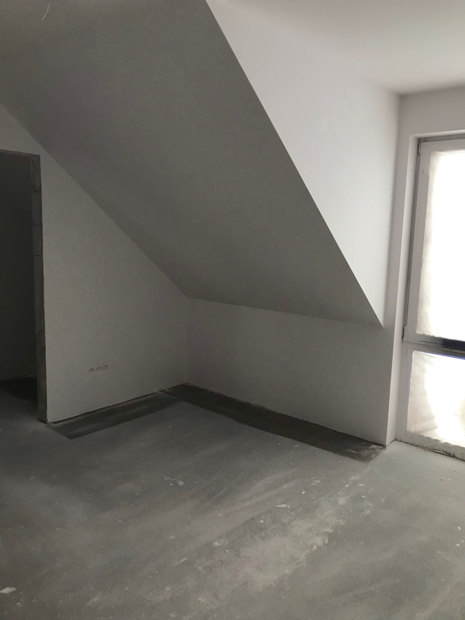 Surowe wnętrze poddasza ze skośnym sufitem, świeżo po gładziowaniu i malowaniu na biało, widoczne ślady po pracach na betonowej podłodze, zabezpieczone okno.