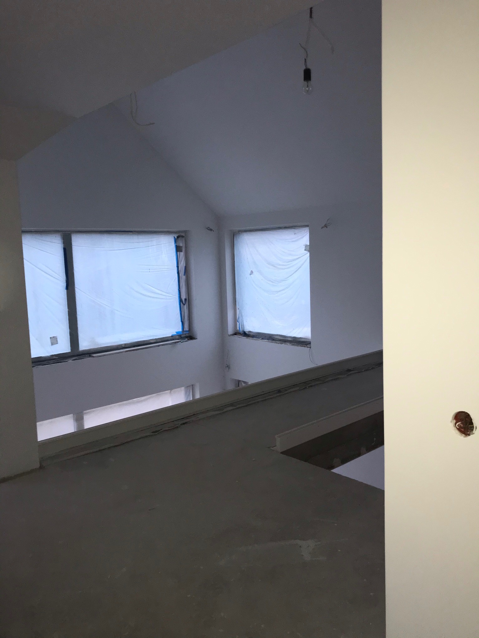 Surowe wnętrze poddasza ze skośnymi ścianami i oknami zabezpieczonymi folią malarską, widoczna surowa podłoga i wystające kable elektryczne.