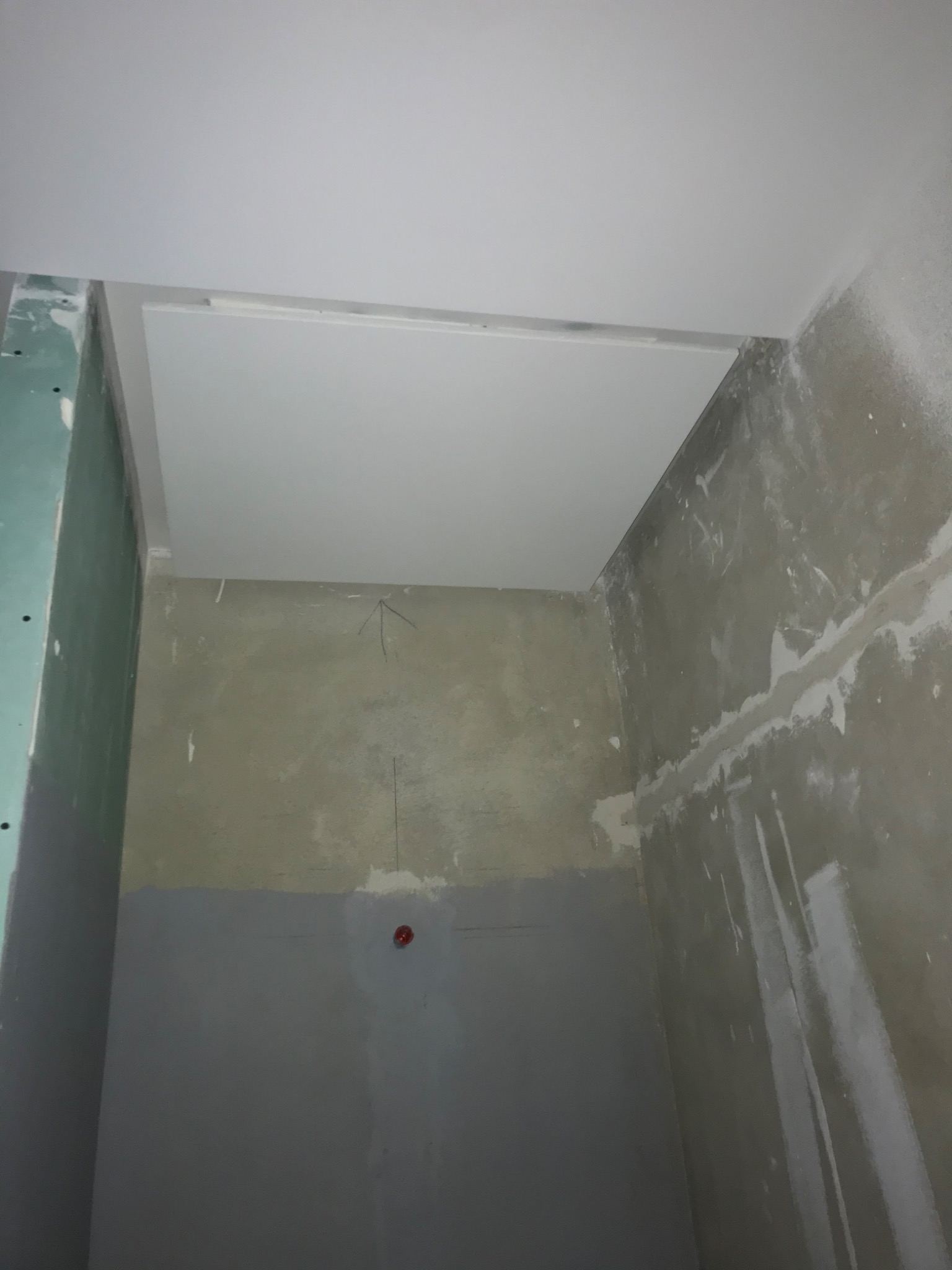 Wnętrze w trakcie remontu: surowa ściana z widocznymi śladami gładzi, częściowo pomalowana na szaro, z zaznaczonym pionem i czerwoną puszką elektryczną, oraz fragment podwieszanego sufitu z płyty...