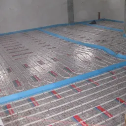 Montaż ogrzewania podłogowego: białe rurki grzewcze ułożone w równych odstępach na siatce z czerwonymi oznaczeniami, zabezpieczone niebieską pianką przy ścianach, widok na surowy betonowy pokój...