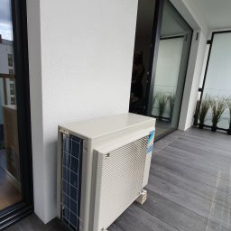 Pompy Ciepła, Klimatyzacje, Fotowoltaika - Zewnętrzna jednostka klimatyzacji Daikin zamontowana na balkonie z szarych desek kompozytowych, widok na przesuwne szklane drzwi z czarnymi ramami i białą ścianę.