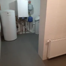 Białe kotły, zasobnik CWU i grzejnik płytowy w kotłowni wyłożonej białymi płytkami, widoczne rury instalacji CO.