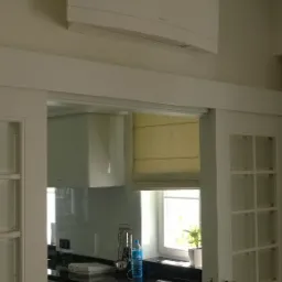 Biały klimatyzator Daikin zamontowany na ścianie w jasnym pomieszczeniu, widok zza białych, przeszklonych drzwi do kuchni z czarnym blatem i kremową roletą w oknie.